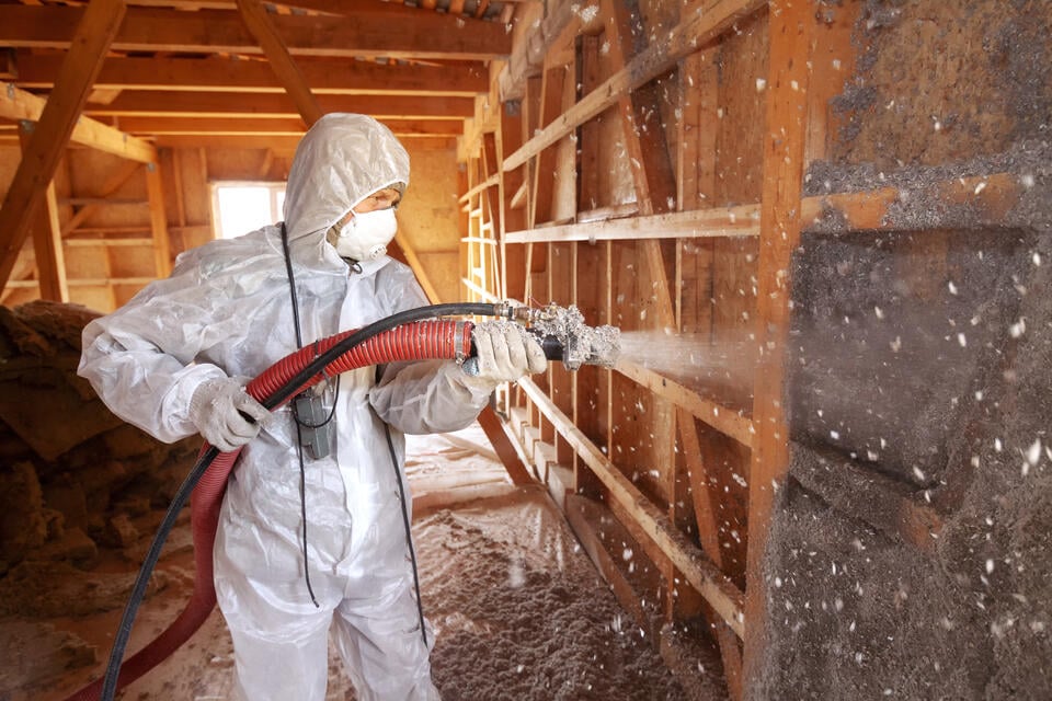 attic insulation cost-technician in protective gear spraying insulation into attic wall