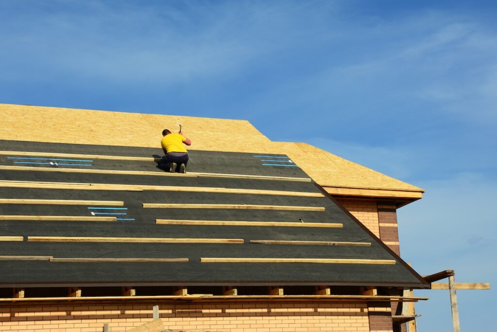 A building constructor is installing roofing underlayment, waterproof, moisture protection on the roof deck of a brick house before covering it with asphalt shingles. Roofing Construction.